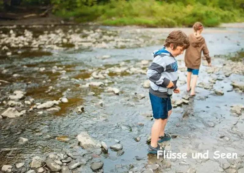 Zwei Kinder mit Shorts und Pullover waten barfuß durch einen flachen, steinigen Fluss im Grünen