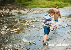 Zwei Kinder mit Shorts und Pullover waten barfuß durch einen flachen, steinigen Fluss im Grünen